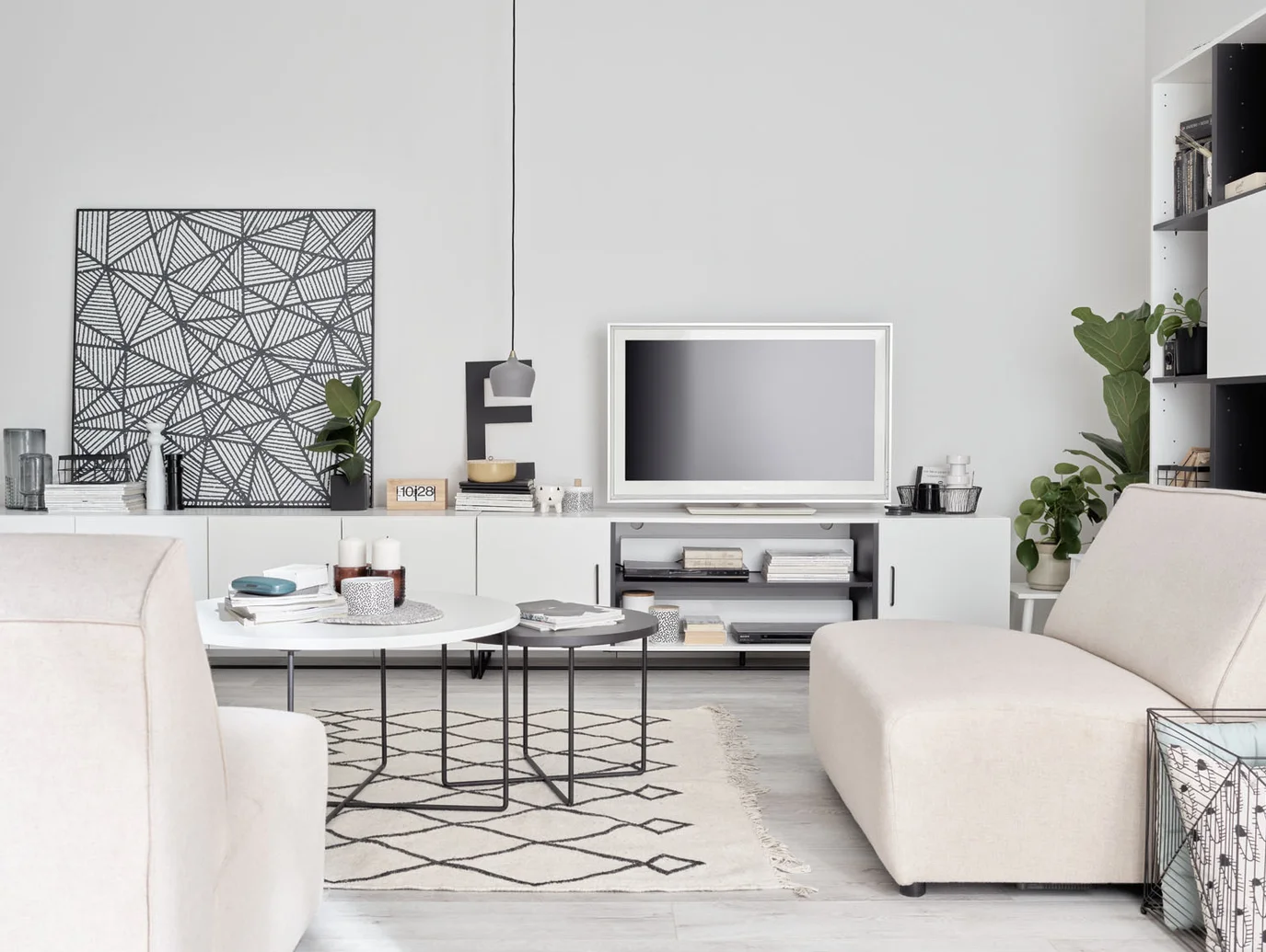Nowoczesny salon w jasnych kolorach z minimalistyczną szafką RTV i telewizorem. Na ścianie geometryczna grafika, a obok rośliny doniczkowe dodające wnętrzu świeżości.